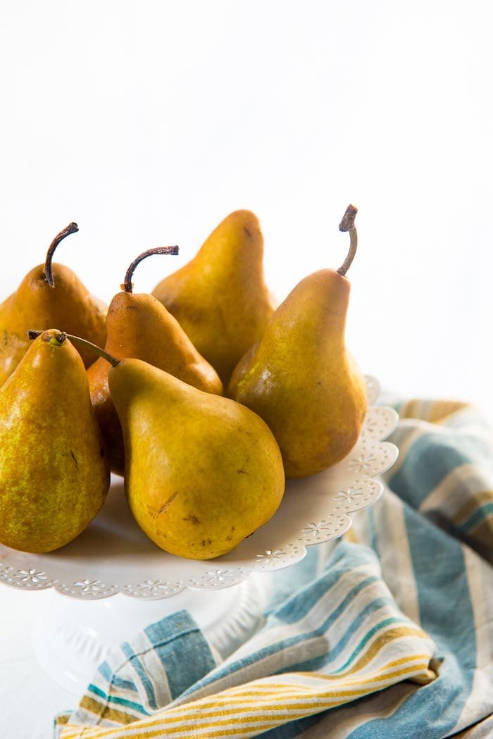 Six Bosc Pears on a white cake stand with a blue green napkin in the background. 