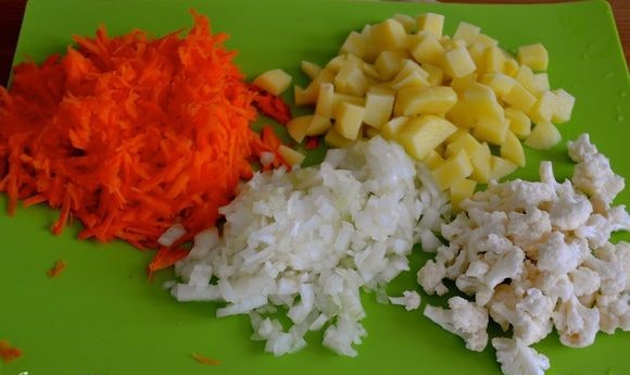нарезанный кубиками картофель, тертая морковь, измельченный репчатый лук и соцветия цветной капусты на зеленой разделочной доске на столе